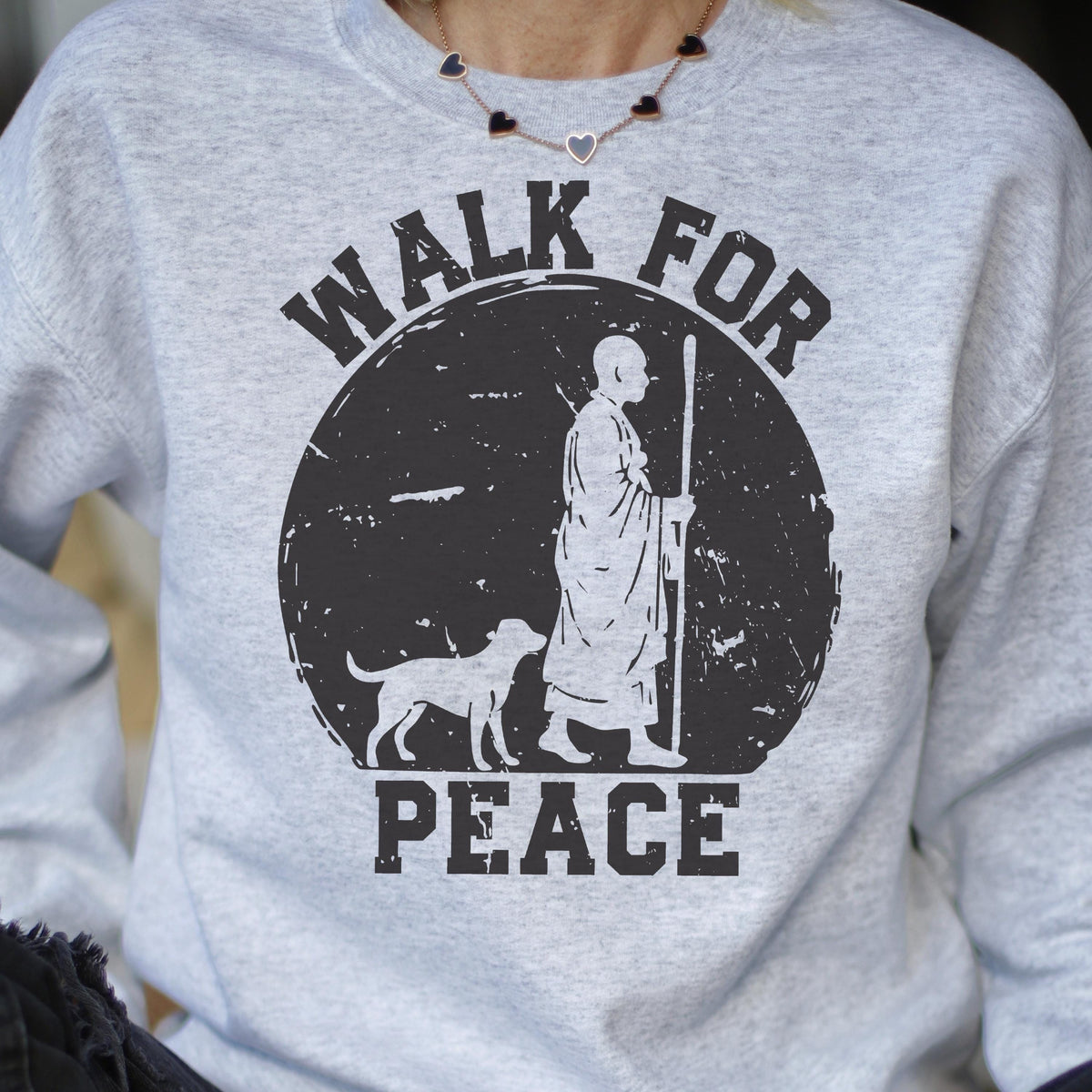 May include: A light gray sweatshirt with a black graphic. The graphic features a figure with a dog, a staff, and the words "WALK FOR PEACE" in a circular design. The sweatshirt has a crew neck.