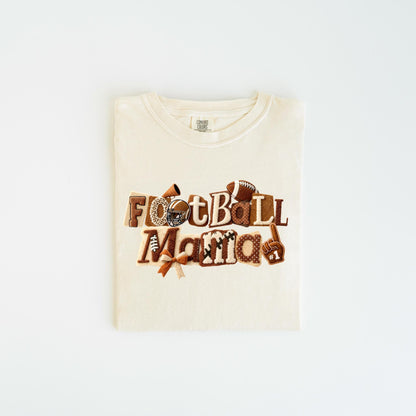 May include: Cream-colored t-shirt with the words "Football Mama" in brown, textured letters. The design includes footballs, a helmet, a megaphone, and a foam finger. The shirt is laid flat on a white surface.