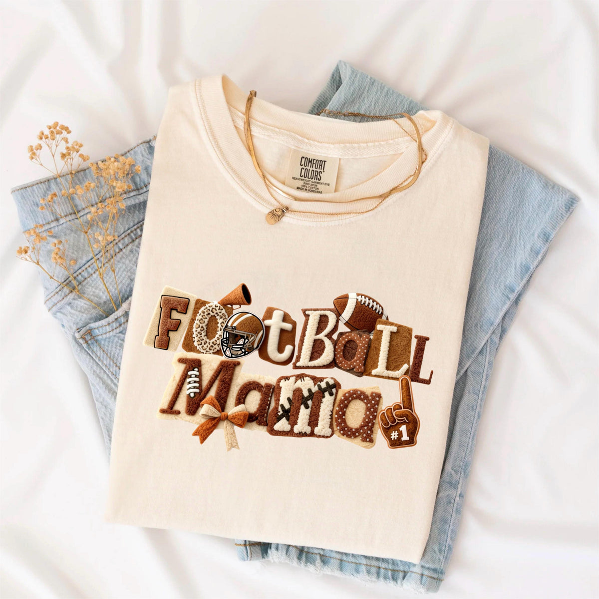 May include: Cream-colored t-shirt with the words "Football Mama" in brown, tan, and white applique. The design includes a football, helmet, and a #1 foam finger. A gold necklace and blue jeans are also in the image.