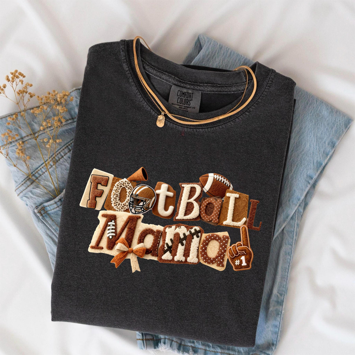 May include: Dark gray Comfort Colors t-shirt with the words "Football Mama" in a decorative design. The design includes a football, helmet, and other football-themed elements. A gold necklace and blue jeans are also visible.