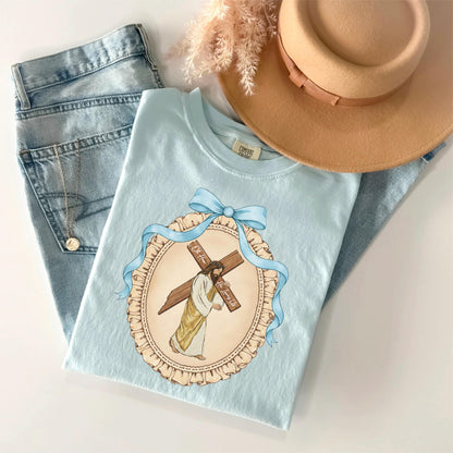 May include: A light blue t-shirt featuring an illustration of Jesus carrying a cross, framed by a blue ribbon and a decorative border. The shirt is paired with light-wash denim jeans and a tan hat. A gold necklace is visible.