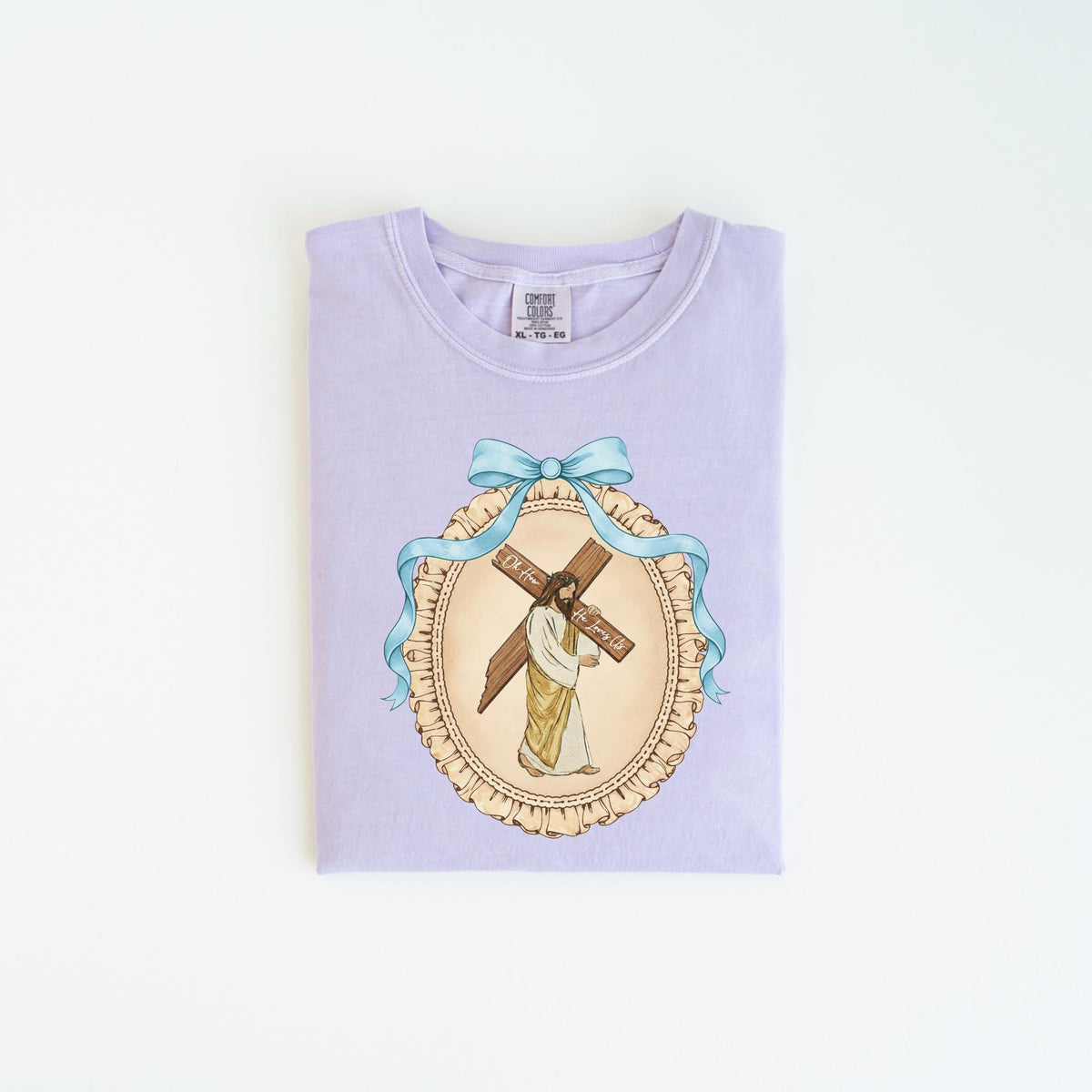 May include: A light purple t-shirt featuring a detailed illustration of Jesus carrying a cross. The image is framed by a light blue ribbon and a decorative oval border. The text on the cross reads "For You, Forever".