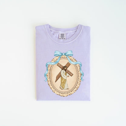 May include: A light purple t-shirt featuring a detailed illustration of Jesus carrying a cross. The image is framed by a light blue ribbon and a decorative oval border. The text on the cross reads "For You, Forever".