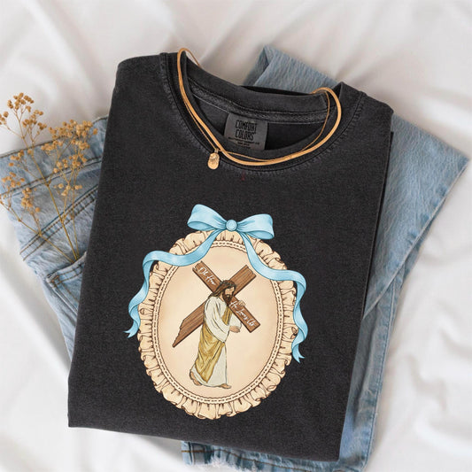 May include: Dark gray Comfort Colors t-shirt featuring a religious graphic of Jesus carrying a cross, with the text "Oh How He Loves Us". The graphic is framed by a light blue ribbon and a decorative border. A gold necklace is draped over the shirt.