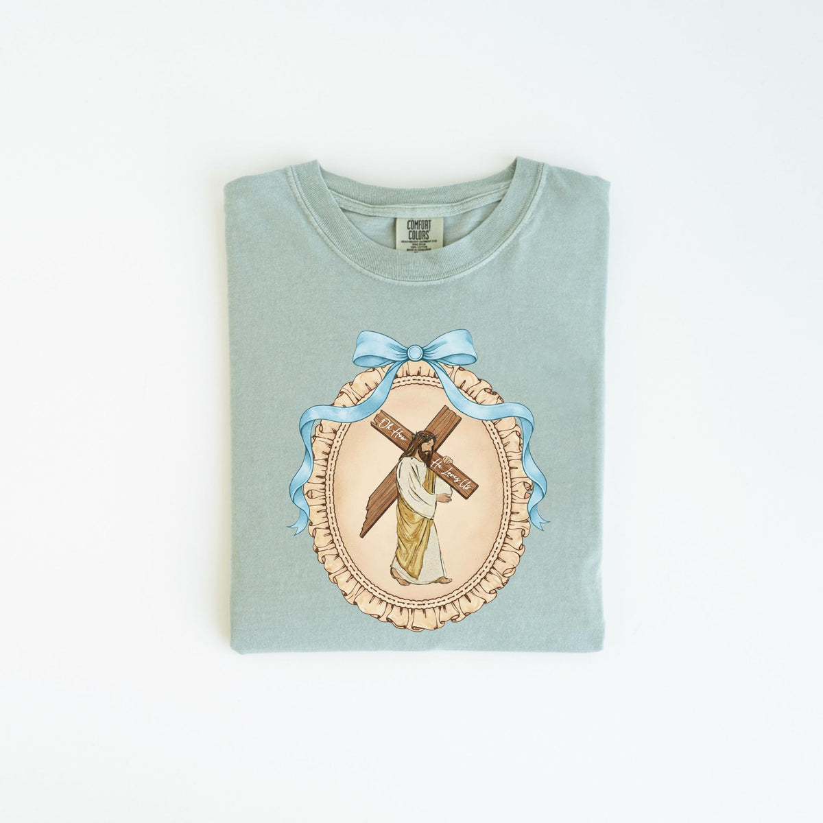 May include: A light green t-shirt featuring a detailed illustration of Jesus carrying a cross. The image is framed by a blue ribbon and a decorative border. The text on the cross reads "Oh For... The Humble".