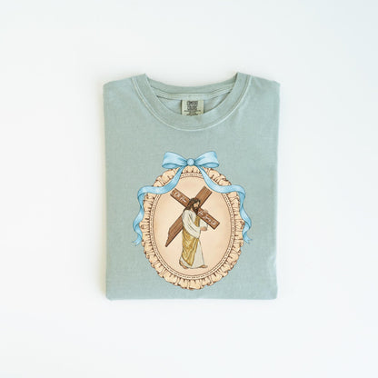 May include: A light green t-shirt featuring a detailed illustration of Jesus carrying a cross. The image is framed by a blue ribbon and a decorative border. The text on the cross reads "Oh For... The Humble".