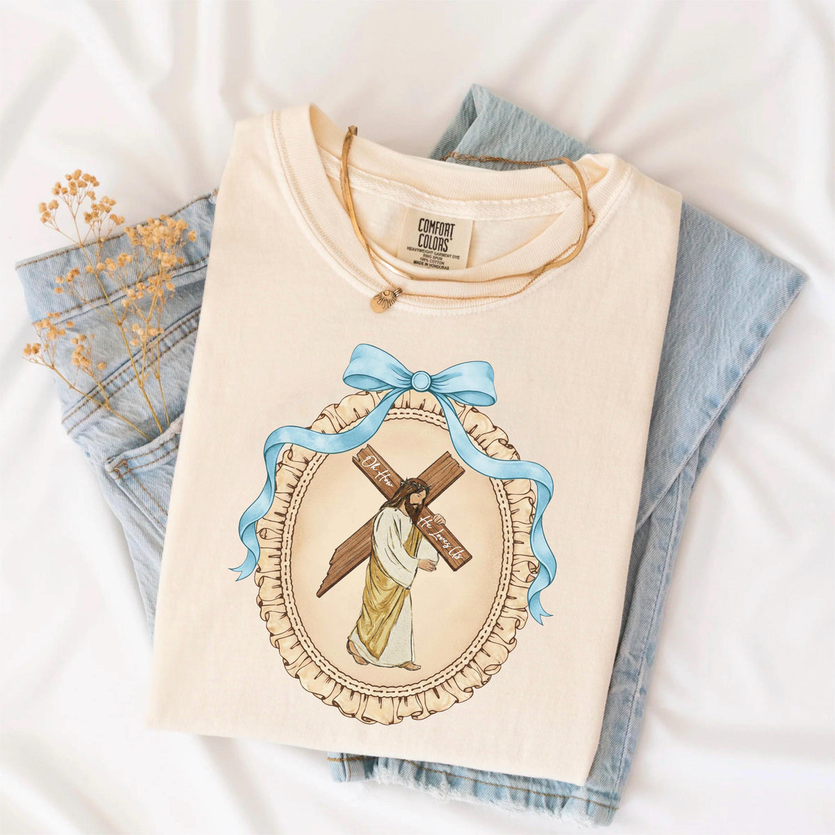 May include: Cream-colored t-shirt featuring a graphic of Jesus carrying a cross, framed by a blue ribbon and a decorative oval. The shirt is folded and placed on a white surface with a pair of blue jeans and a gold necklace.