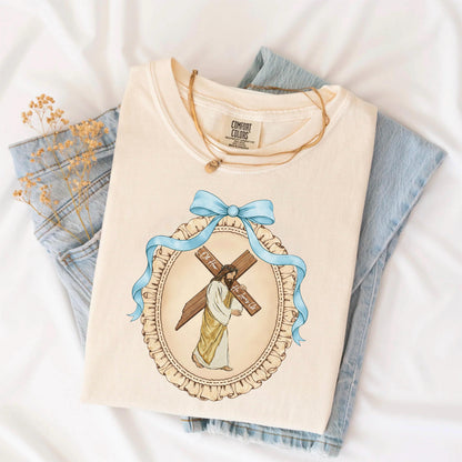 May include: Cream-colored t-shirt featuring a graphic of Jesus carrying a cross, framed by a blue ribbon and a decorative oval. The shirt is folded and placed on a white surface with a pair of blue jeans and a gold necklace.