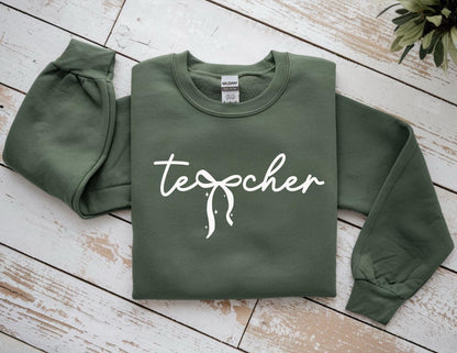 May include: An olive green sweatshirt with the word "teacher" in white script across the front. The sweatshirt has long sleeves and a crew neck. The text has a bow design.