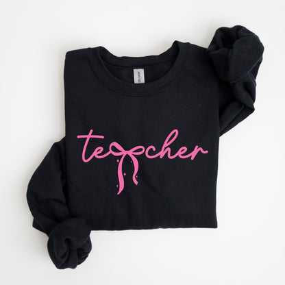 May include: Black crewneck sweatshirt with the word "teacher" in pink script. The word "teacher" has a pink bow design in the center. The sweatshirt is folded neatly on a white surface.