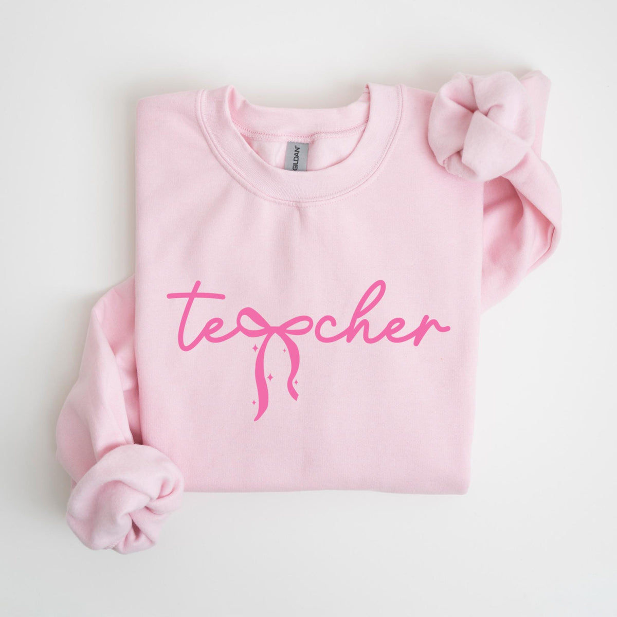 May include: A light pink sweatshirt with the word "teacher" in a cursive font, with a bow graphic. The sleeves are folded. The sweatshirt is made of a soft material.