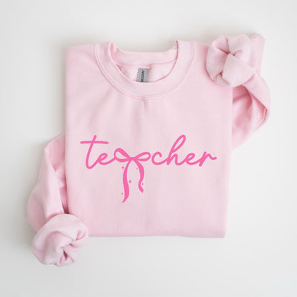 May include: A light pink sweatshirt with the word "teacher" in a cursive font, with a bow graphic. The sleeves are folded. The sweatshirt is made of a soft material.