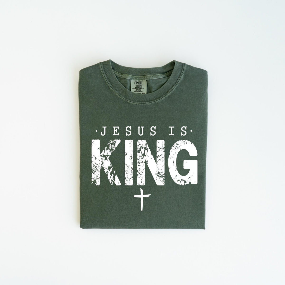 May include: Dark green t-shirt with the text "JESUS IS KING" in distressed white lettering and a white cross below. The shirt is folded and laid flat on a white surface.