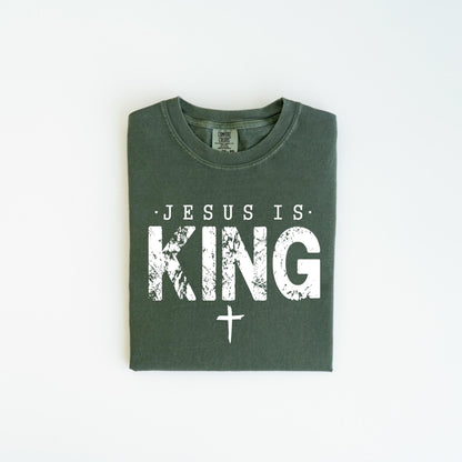 May include: Dark green t-shirt with the text "JESUS IS KING" in distressed white lettering and a white cross below. The shirt is folded and laid flat on a white surface.