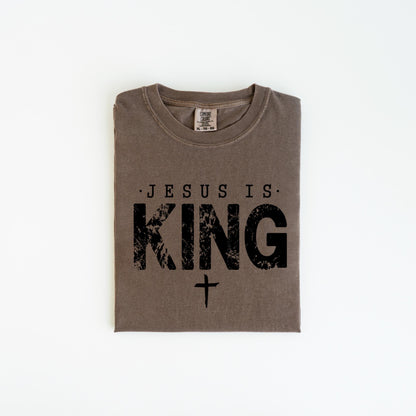 May include: A folded, brown t-shirt with the text "JESUS IS KING" in black, distressed lettering, and a small cross below. The shirt is made of a soft material and has a crew neck.