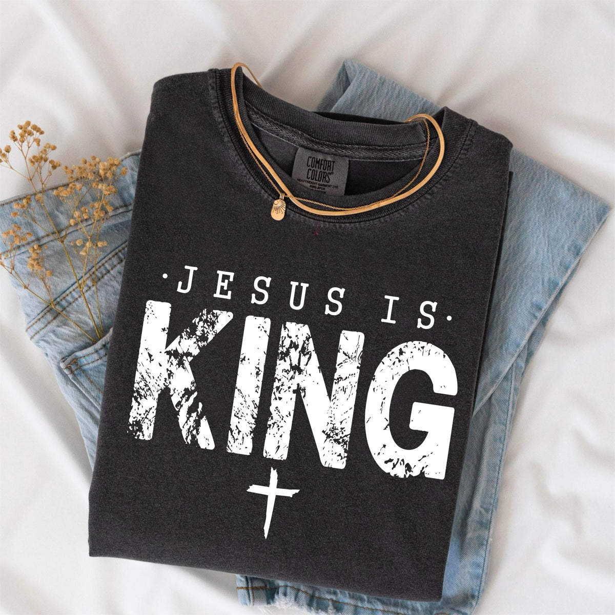 May include: Dark gray t-shirt with the text "JESUS IS KING" in distressed white lettering and a white cross. The shirt is folded on top of a pair of blue jeans. A gold necklace is draped on the shirt.