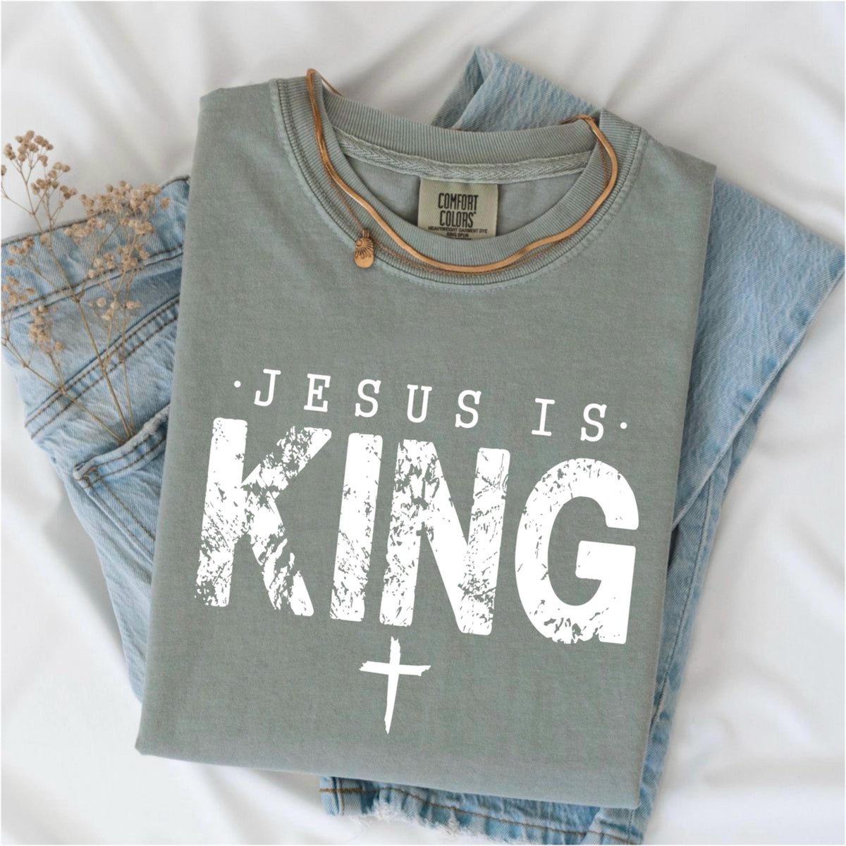 May include: Sage green t-shirt with the text "JESUS IS KING" in distressed white lettering and a white cross. The shirt is folded on top of blue jeans, with a brown necklace and dried flowers. The shirt has a Comfort Colors tag.