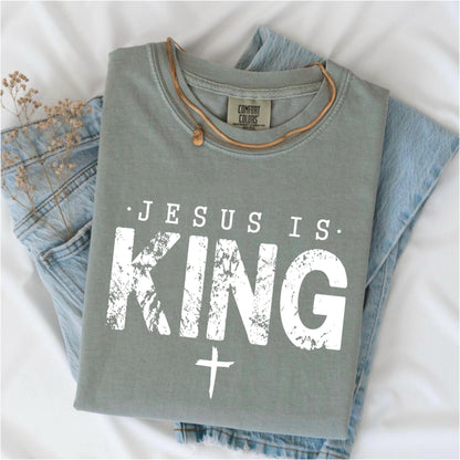 May include: Sage green t-shirt with the text "JESUS IS KING" in distressed white lettering and a white cross. The shirt is folded on top of blue jeans, with a brown necklace and dried flowers. The shirt has a Comfort Colors tag.