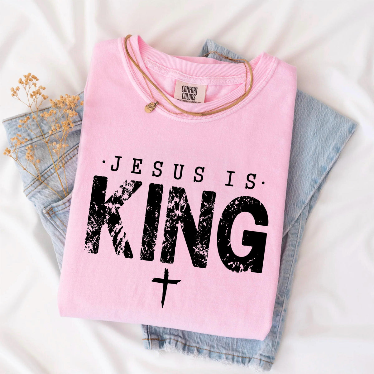 May include: A light pink t-shirt with the text "JESUS IS KING" in black, along with a cross symbol. The shirt is folded and placed on top of a pair of blue jeans. A gold necklace is draped across the shirt.