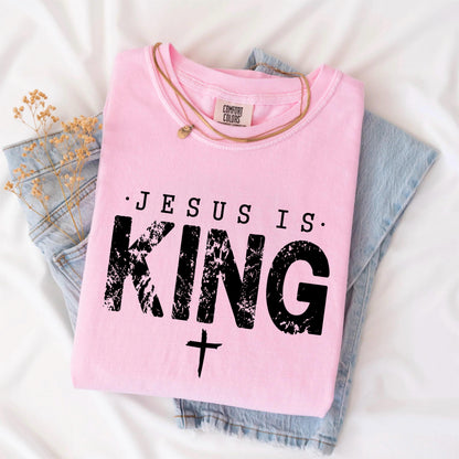 May include: A light pink t-shirt with the text "JESUS IS KING" in black, along with a cross symbol. The shirt is folded and placed on top of a pair of blue jeans. A gold necklace is draped across the shirt.