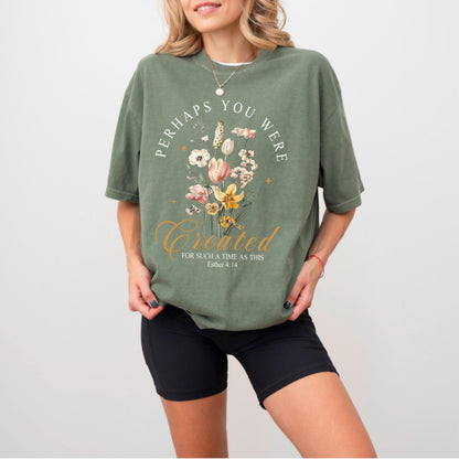 May include: Olive green t-shirt with the text "PERHAPS YOU WERE CREATED FOR SUCH A TIME AS THIS" and a floral design. The design includes pink, yellow, and white flowers. The shirt is paired with black shorts.