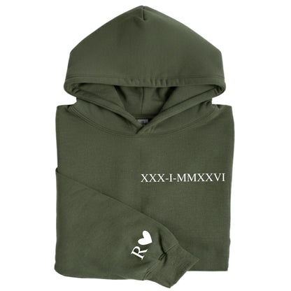May include: An olive green hooded sweatshirt featuring the white text XXX-I-MMXXVI printed on the chest. The left sleeve displays a white letter R followed by a small white heart symbol. The garment has a hood and long sleeves with ribbed cuffs.