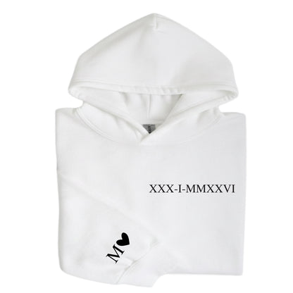 May include: A white hooded sweatshirt featuring black text on the chest that reads XXX-I-MMXXVI. The left sleeve cuff displays the letter M followed by a small black heart symbol. The garment has a standard hood, long sleeves, and a front pocket.