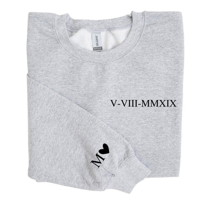 May include: A light gray crewneck sweatshirt featuring the black Roman numeral date V-VIII-MMXIX printed on the chest. The left sleeve displays the letter M followed by a small black heart symbol. The garment has a standard ribbed collar and cuffs.