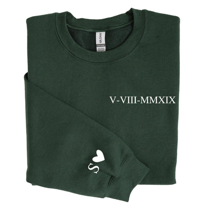 May include: A dark forest green crewneck sweatshirt featuring the white Roman numeral date V-VIII-MMXIX printed on the chest and a white letter S followed by a heart symbol printed on the left sleeve cuff. The garment has a standard ribbed neckline.