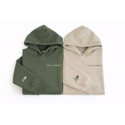 May include: Two hooded sweatshirts, one olive green and one beige. Each has a hood, long sleeves, and the Roman numeral date "XXIII IX MMXVII" printed on the chest. The left sleeve of each sweatshirt has a letter and a heart.