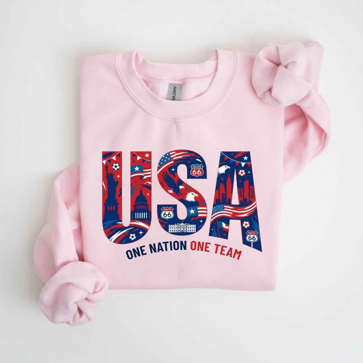 May include: A light pink sweatshirt with the word "USA" in red, white, and blue, filled with patriotic imagery. The text "ONE NATION ONE TEAM" is below the word "USA".