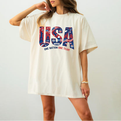 May include: Cream-colored t-shirt with the word "USA" in red and blue, featuring patriotic imagery. The text "ONE NATION ONE TEAM" is below the word "USA". The shirt has short sleeves and a relaxed fit.
