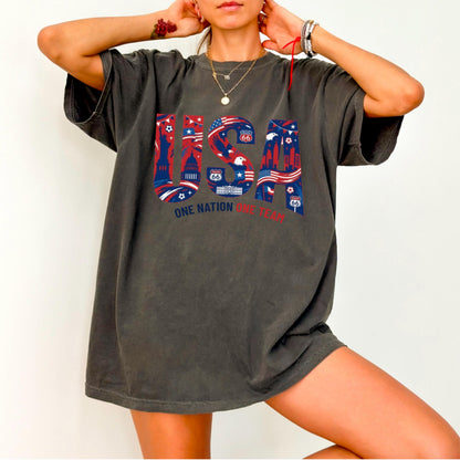 May include: Dark grey t-shirt with a USA graphic in red, white, and blue. The graphic includes the text "USA", the Route 66 sign, and the words "ONE NATION ONE TEAM". The shirt has short sleeves and a relaxed fit.