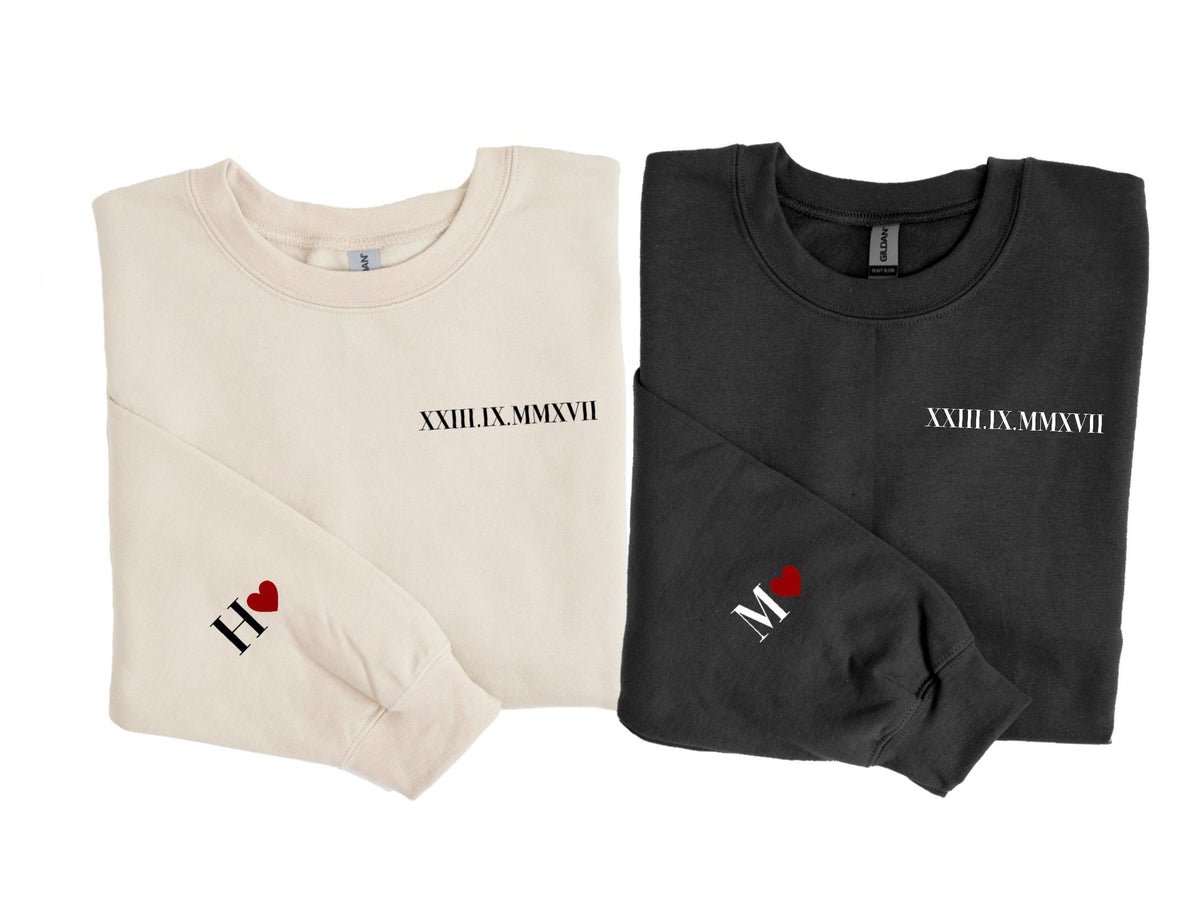 May include: Two long-sleeved sweatshirts, one cream and one black, are displayed. Each has the Roman numeral date "XXIII.IX.MMXVII" printed on the chest. The cream sweatshirt has "H" and a heart on the sleeve, while the black one has "M" and a heart.