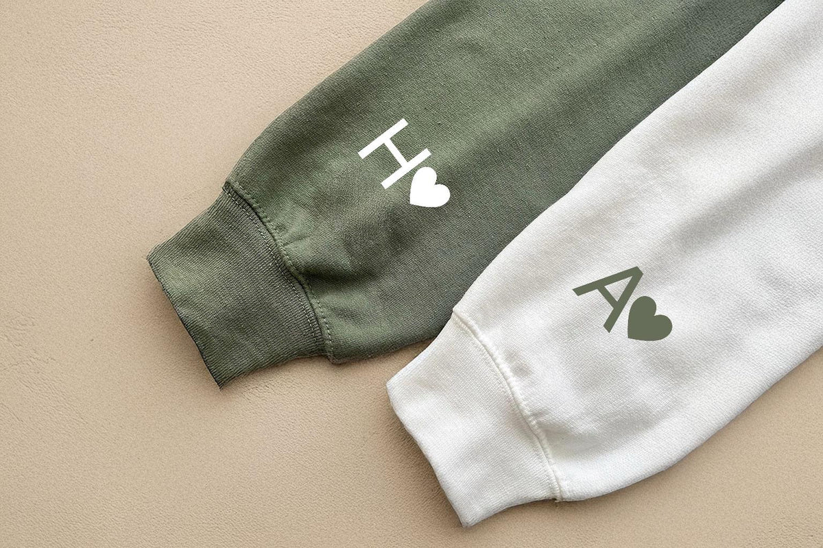 May include: Close-up of two sweatshirt sleeves, one olive green and one white. The olive sleeve has a white "H" and heart, the white sleeve has a green "A" and heart. The sleeves are on a tan surface.