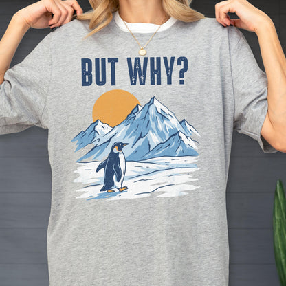 May include: A gray t-shirt featuring a graphic of a penguin standing in front of snow-covered mountains and a yellow sun. The text "BUT WHY?" is printed above the graphic in dark blue letters. The shirt is made of a soft material.