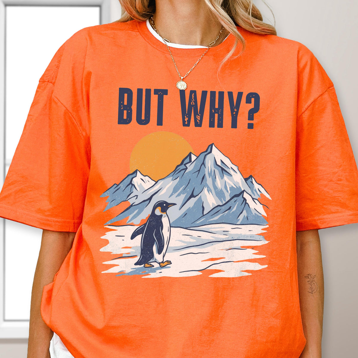 May include: Orange t-shirt with the text "BUT WHY?" in navy blue. The graphic features a penguin standing on snow in front of mountains and a setting sun. The shirt is a crew neck style.