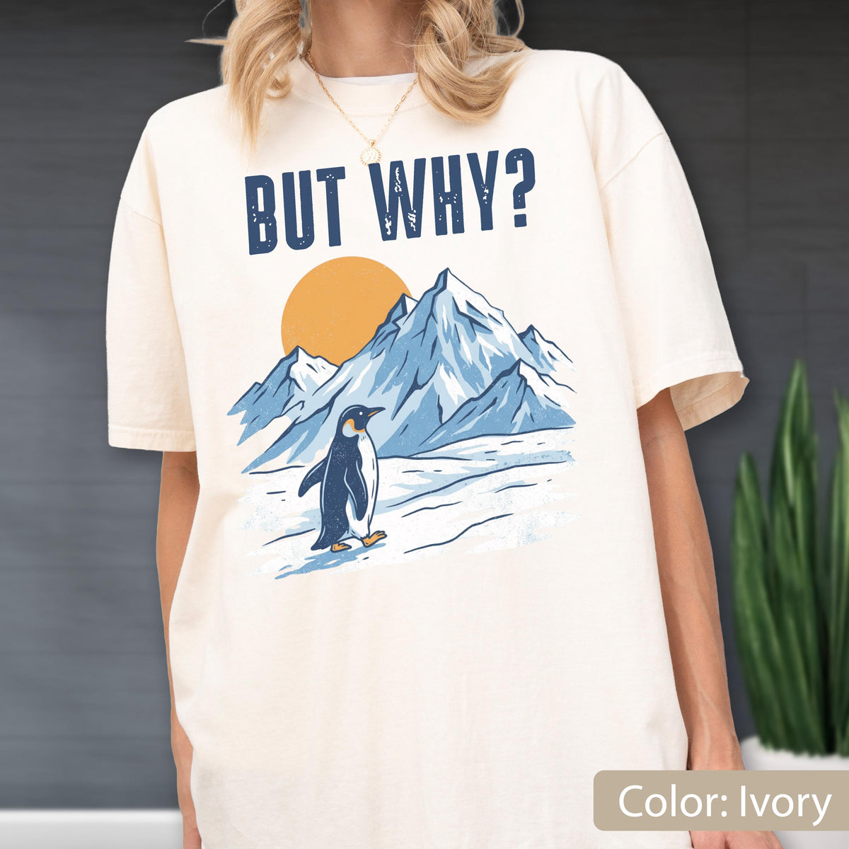 May include: Ivory-colored t-shirt featuring a graphic of a penguin walking in front of mountains and a sun. The text "BUT WHY?" is printed above the graphic. The shirt is a relaxed fit.