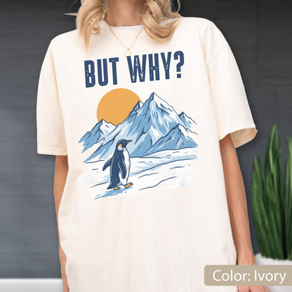 May include: Ivory-colored t-shirt featuring a graphic of a penguin walking in front of mountains and a sun. The text "BUT WHY?" is printed above the graphic. The shirt is a relaxed fit.