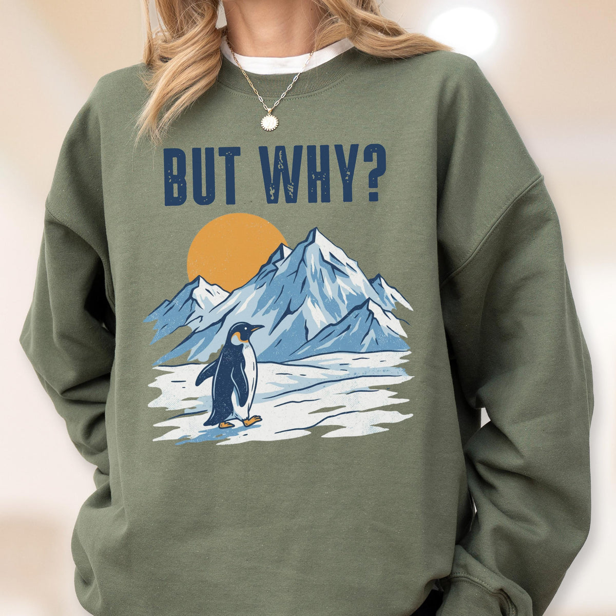 May include: Olive green sweatshirt with a graphic of a penguin standing in front of snow-covered mountains and a setting sun. The text "BUT WHY?" is printed above the graphic in blue letters. The sweatshirt is made of a soft material.