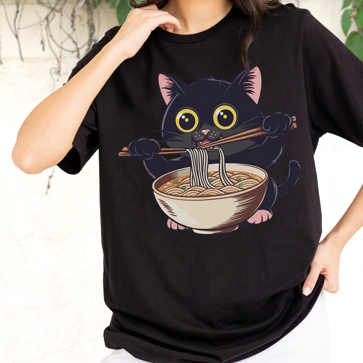May include: Black t-shirt featuring a cartoon black cat with large yellow eyes, holding chopsticks and eating noodles from a bowl. The cat has pink inner ears and paws. The design is centered on the shirt.