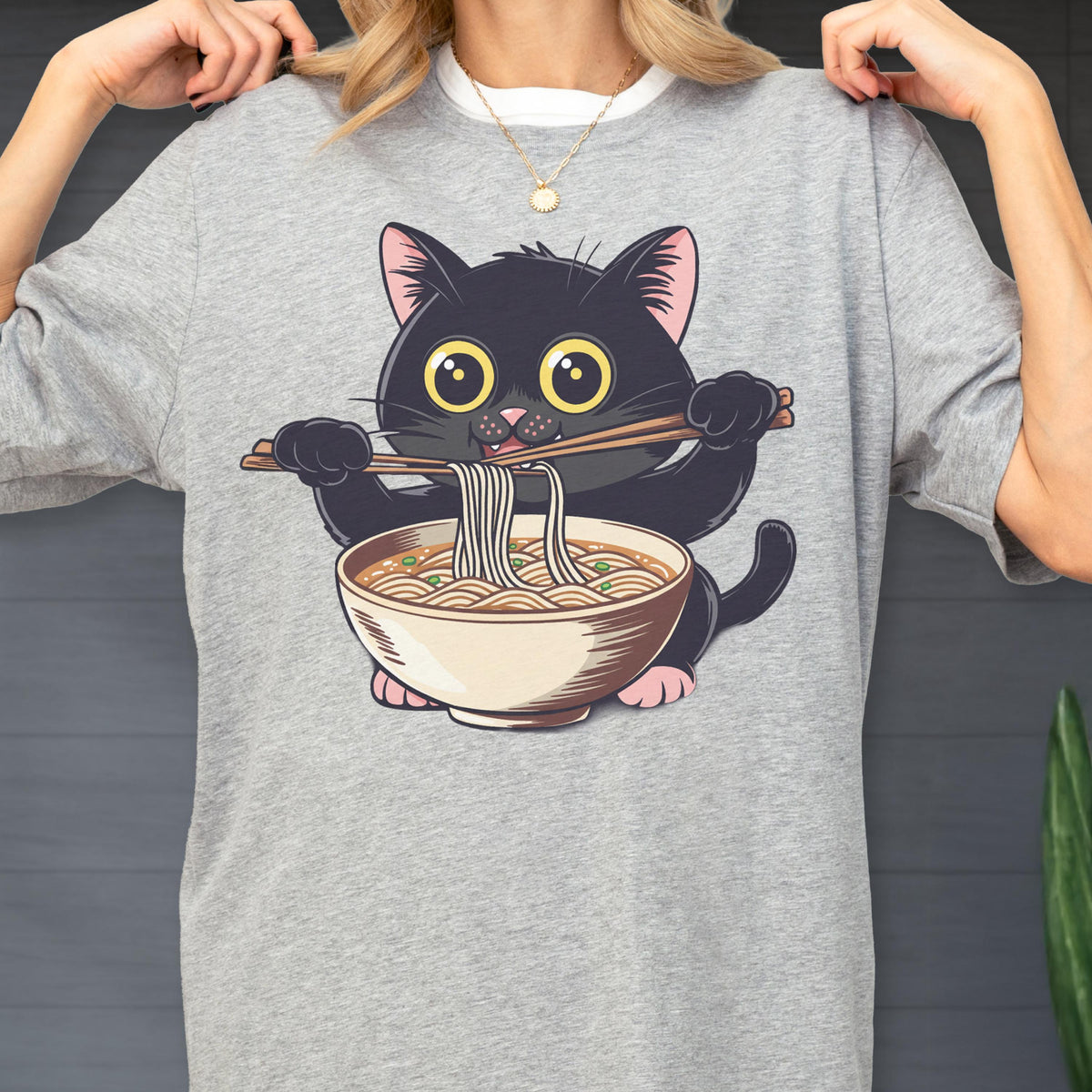 May include: A gray t-shirt featuring a cartoon black cat holding chopsticks and a bowl of ramen. The cat has large yellow eyes and pink ears and paws. The ramen bowl is beige with brown broth and green garnish.