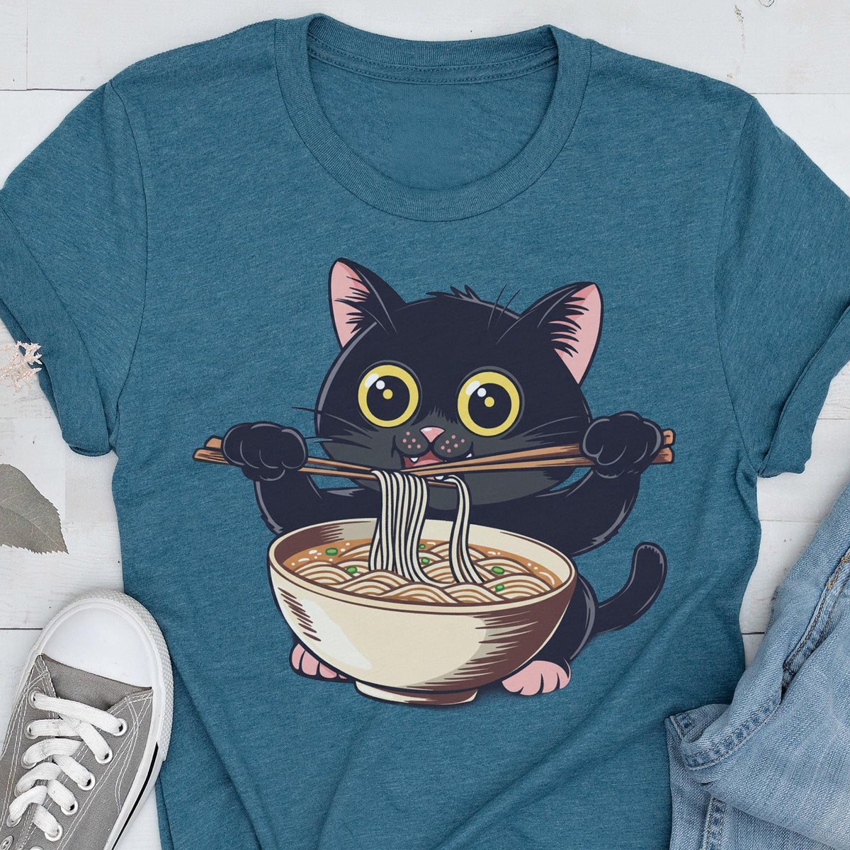 May include: Teal t-shirt featuring a cartoon black cat with large yellow eyes, holding chopsticks with noodles over a bowl of ramen. The cat has pink ears and paws. The design is centered on the shirt.