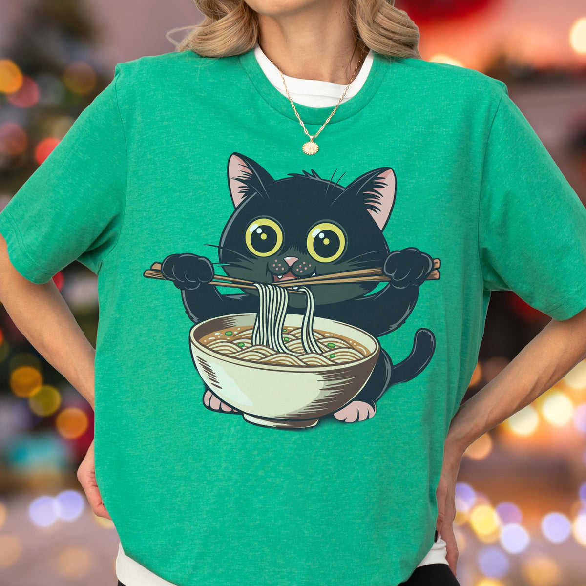 May include: Green t-shirt featuring a cartoon black cat eating ramen noodles with chopsticks. The cat has large yellow eyes and is holding the chopsticks with its paws. The ramen bowl is white with a brown rim and contains noodles and broth.