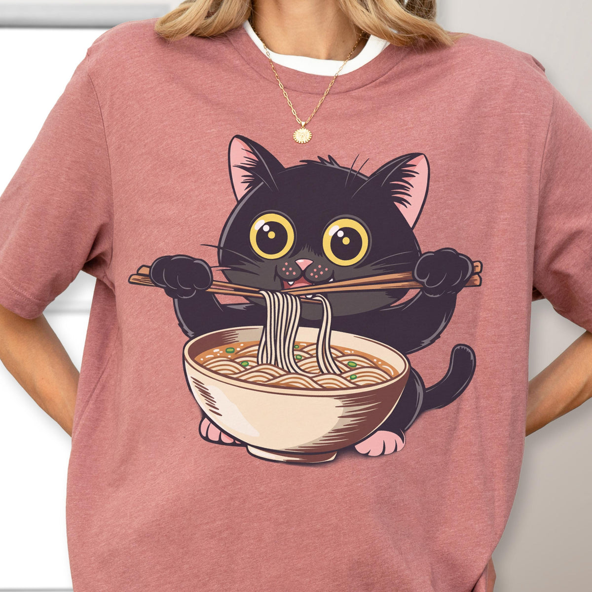 May include: A dusty rose t-shirt featuring a cartoon black cat holding chopsticks with noodles over a bowl of ramen. The cat has large yellow eyes and pink paws. The shirt is a casual style.