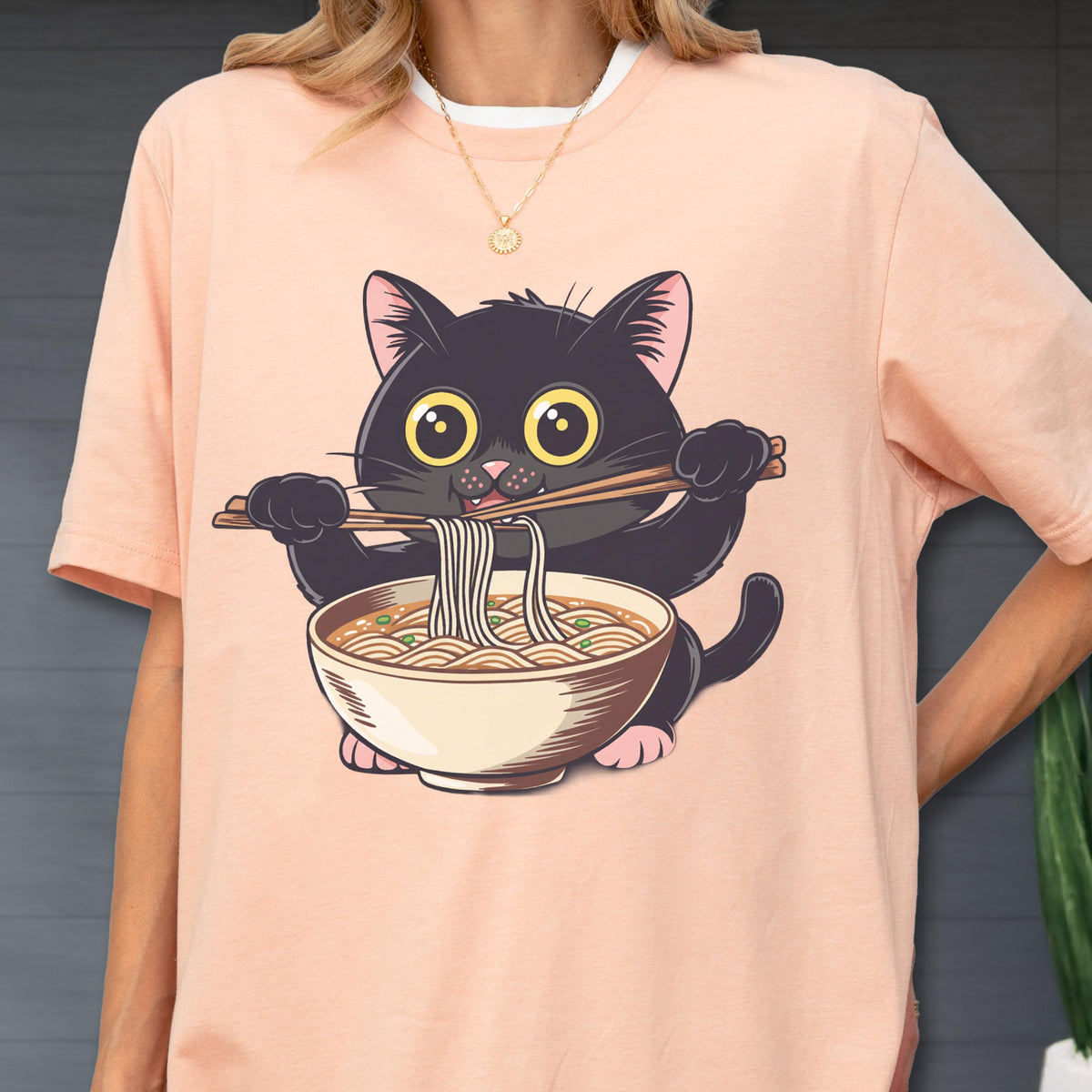 May include: A light pink t-shirt featuring a cartoon black cat eating ramen noodles with chopsticks. The cat has large yellow eyes and is holding a bowl of noodles. The shirt has a white collar.