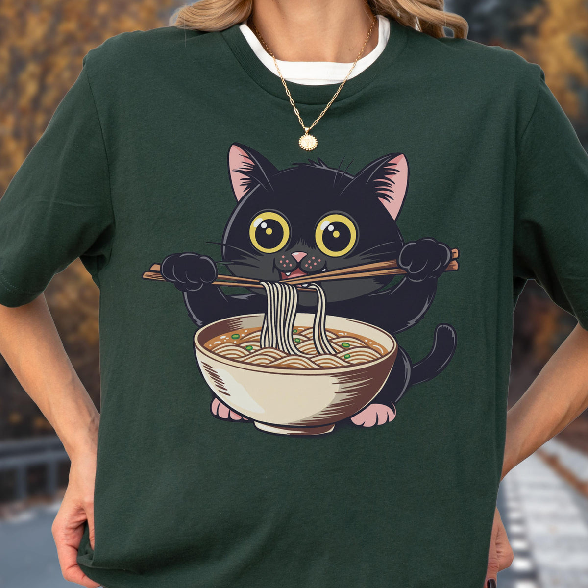 May include: Dark green t-shirt featuring a cartoon black cat eating ramen noodles with chopsticks. The cat has large yellow eyes and pink ears and paws. The ramen bowl is beige with a brown broth and noodles.