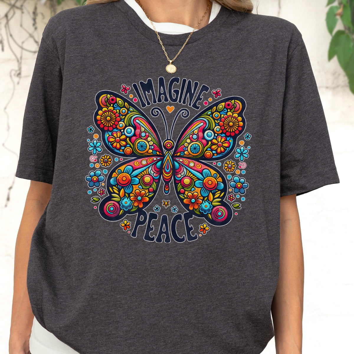 May include: A charcoal gray short-sleeve t-shirt featuring a large, colorful butterfly graphic with intricate floral patterns in shades of red, orange, yellow, and blue. The text "IMAGINE" is printed above the butterfly and "PEACE" below it.