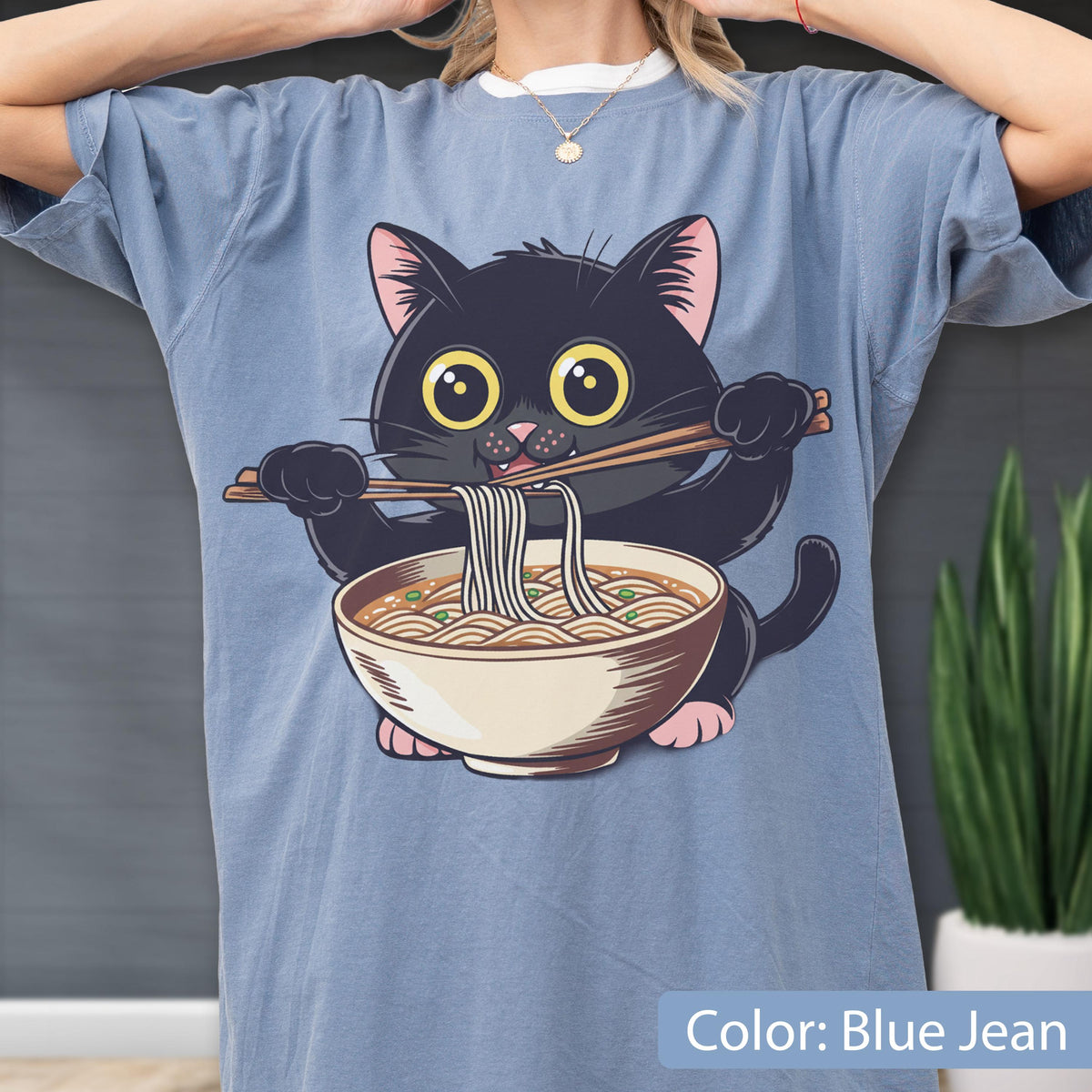 May include: Blue jean colored t-shirt featuring a cartoon black cat eating ramen noodles from a bowl with chopsticks. The cat has large yellow eyes and pink accents. The text "Color: Blue Jean" is at the bottom of the image.