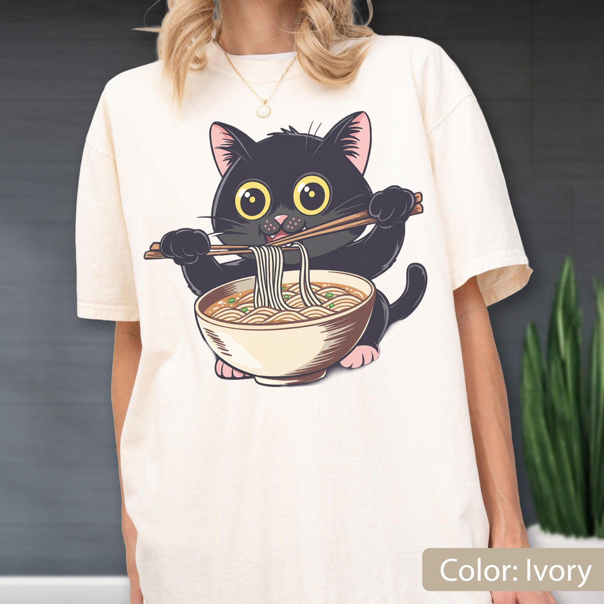 May include: Ivory-colored t-shirt featuring a cartoon black cat eating ramen noodles with chopsticks. The cat has large yellow eyes and pink paws. The ramen bowl is beige with brown noodles. The text "Color: Ivory" is at the bottom.